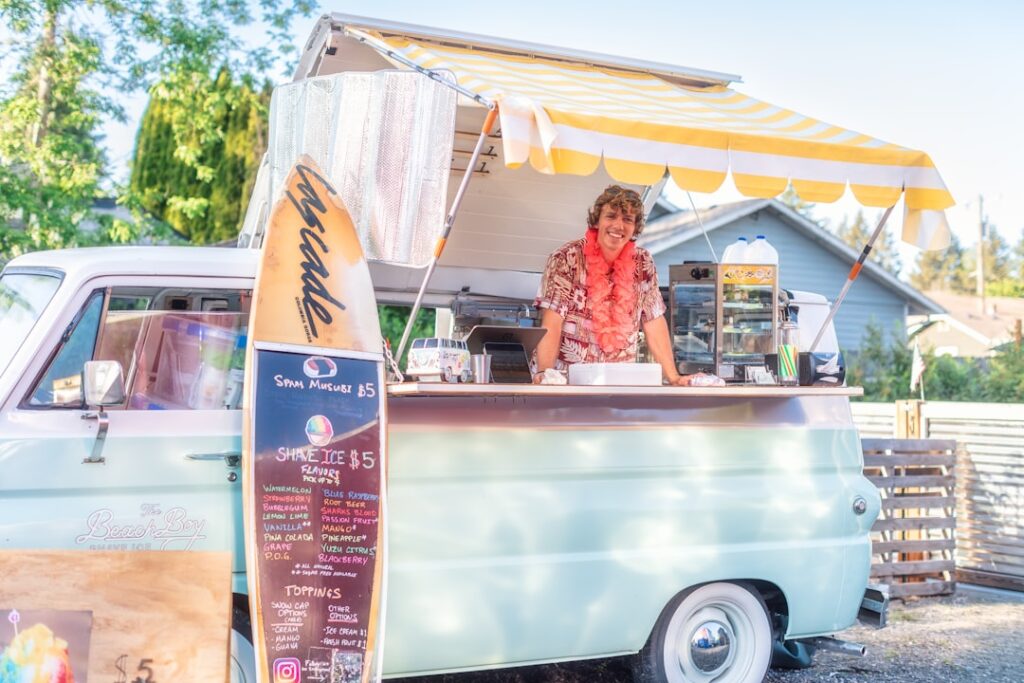 a woman standing behind a food truck with a surfboard on the back of it