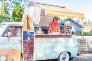 a woman standing behind a food truck with a surfboard on the back of it